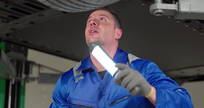 Mechanic inspecting car underbody with work light under vehicle on lift in modern garage