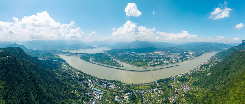 A wide aerial view shows the majestic Three Gorges Dam spanning across the large river surrounded by green mountains and blue sky on a sunny summer day