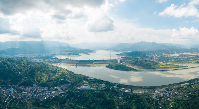 A wide aerial view of the Three Gorges Dam spanning across the large river with surrounding green mountains under a bright cloudy sky during the day