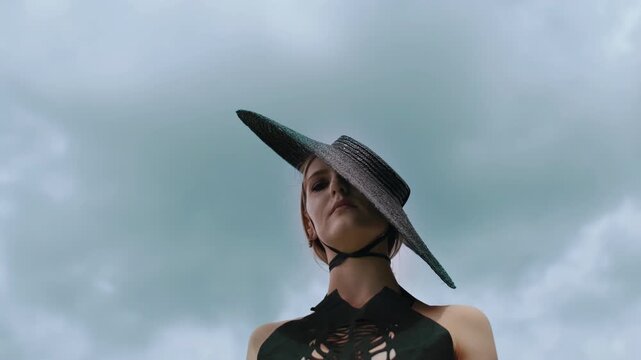 Extreme low-angle view of adult woman in pointed wide-brim straw hat and dark strappy cutout top slowly adjusting head position against expansive overcast sky, creating moody fashion mood and