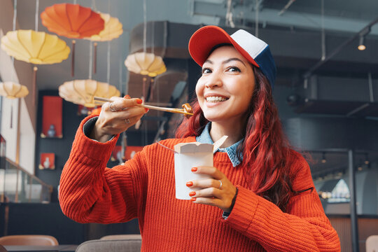 Young woman with red hair wearing a cap happily eating Asian cuisine from a cardboard box using chopsticks indoors