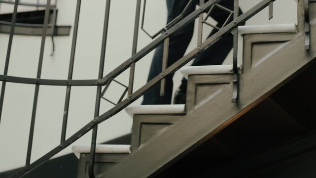 Cropped interior motion shot showing person ascending staircase, visible by cropped legs and shoes on consecutive marble-topped concrete steps behind vertical black metal balusters