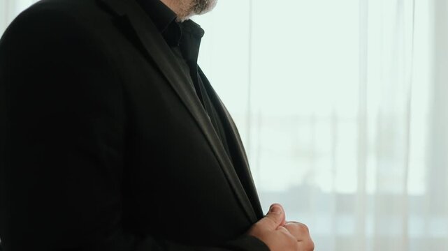 Side view shows person adjusting dark tailored suit jacket at chest with both hands, partial lower face and trimmed beard visible, soft daylight from window creating contrast, polished business mood