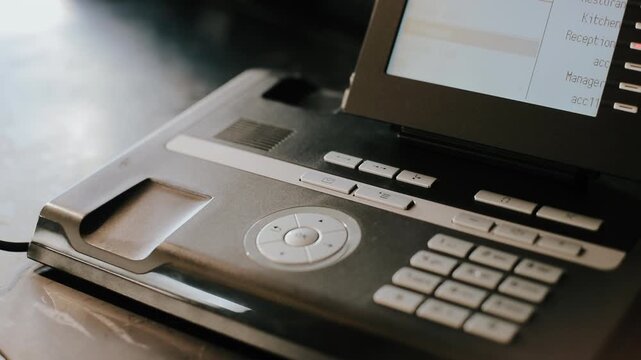 Close view of office desk telephone showing handset lifted and returned by hand, with keypad detail, curved cord, warm directional light, subtle reflections, and calm workplace communication setting