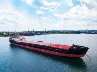 Fototapeta premium Large cargo ship navigates through calm waters near port on a sunny day with scattered clouds and cranes in the background