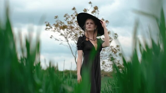 Young woman in flowing black dress steadies wide-brim black hat with sheer net veil, turning from profile toward three-quarter view with poised, contemplative expression amid tall grass and soft