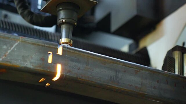 Laser cutting nozzle machining rectangular steel tube with bright sparks and molten slag at cut edge inside industrial workshop, showing precise metal fabrication and heat glow