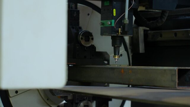 Industrial laser cutting head producing bright sparks while slicing rectangular steel tube inside fabrication machine with precise metalwork detail and glowing contact point
