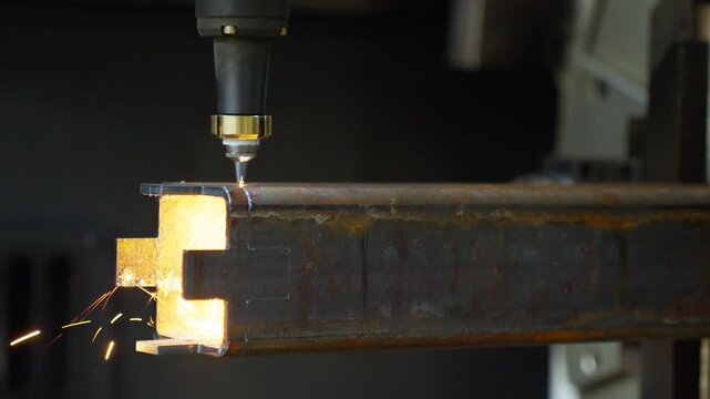 Industrial laser cutting nozzle creating bright molten opening and sparks on rusted rectangular steel tube inside workshop with precise metal removal and glowing cut edge