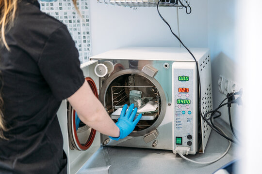 Medical worker in glove opens door of fractionated vacuum autoclave to load dental tools for sanitization. Autoclave sterilizer machine, dental sterilization equipment, medical autoclave technology.