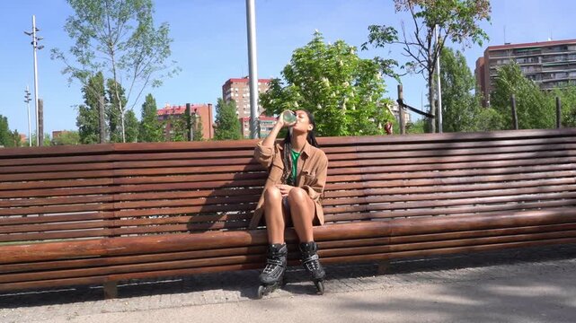 Young woman with inline skates drinking water on bench