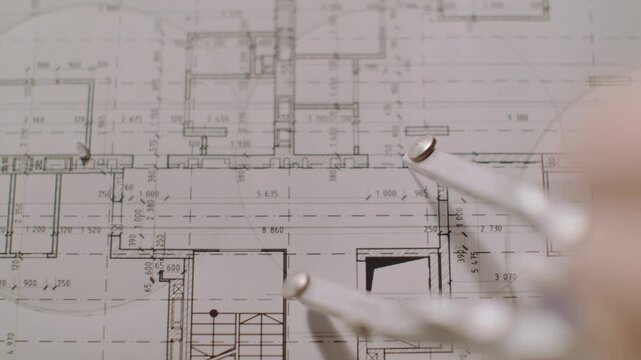 Soft blurred hand and metal drafting tools pass over detailed architectural floor plan, with room outlines, measurements, and technical linework visible beneath warm neutral light in calm