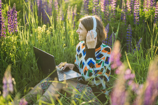 Smiling woman uses laptop and headphone in sunny flower field