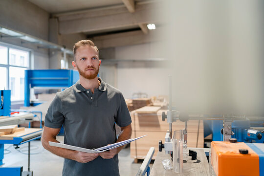 Portrait of carpenter inspecting machines in production hall with ring binder in hands
