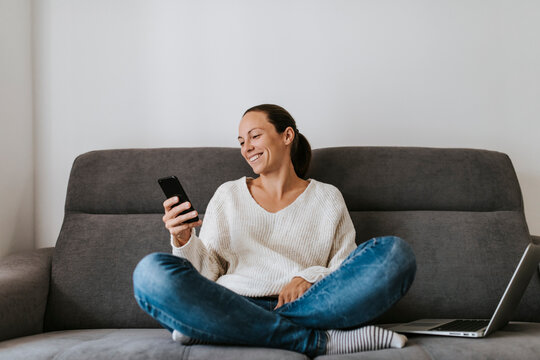 Smiling woman using smart phone while sitting with laptop on sofa