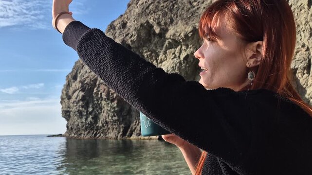 Woman drinking coffee by the sea reaching for sunlight, capturing a calm moment with an eye-level medium shot.
