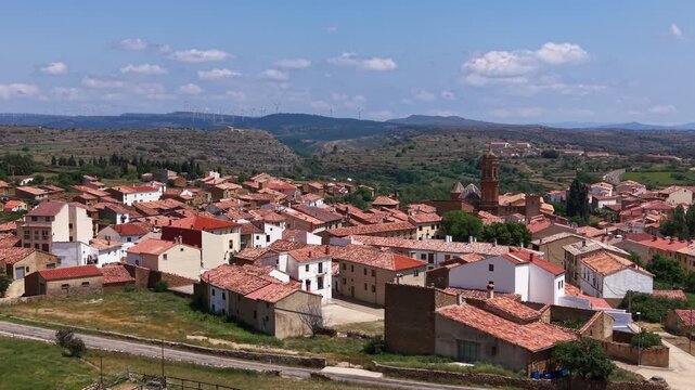 Panoramic view of La Iglesuela del Cid, a medieval village in Maestrazgo, Teruel, Spain, featuring terracotta rooftops, stone church bell tower, and wind turbines on distant ridge