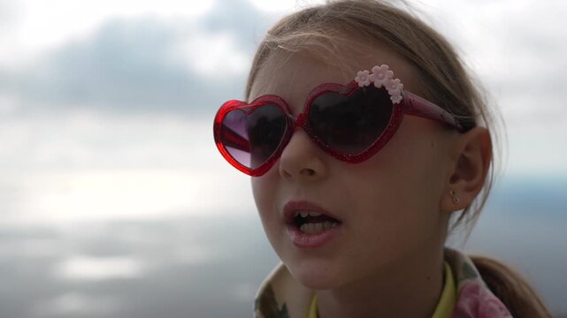 Child sunglasses heart-shaped. Close-up portrait of a cute young girl wearing red novelty glasses outside on a sunny, windy day near the water.