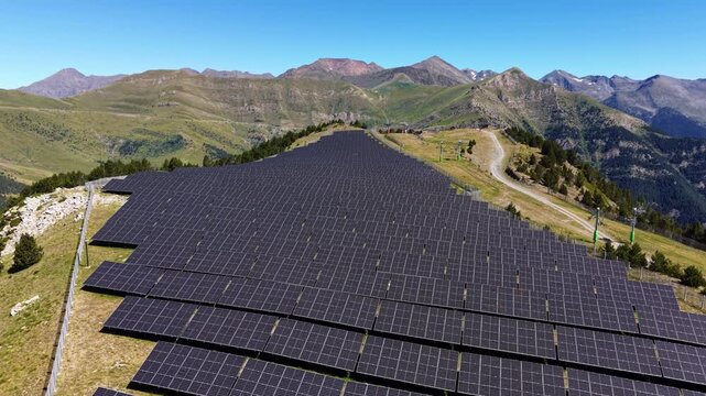 Dense rows of solar panels covering a mountain ridge in Andorra, with ski lift infrastructure and layered Pyrenean peaks under a clear blue sky in the background
