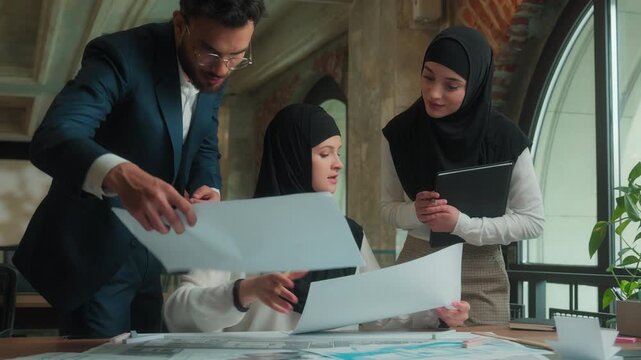 Arabian buiness team Muslim designer architect woman in hijab drawing blueprints two colleagues businessman businesswoman coming in office checking review architecture project plan three co-workers