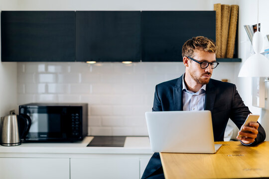 Young businessman using laptop and cell phone in kitchen