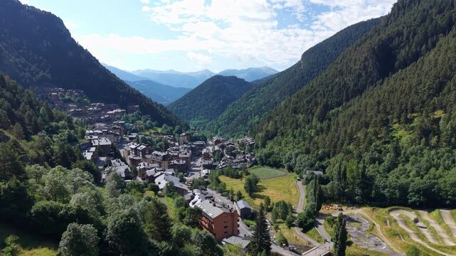 Elevated view of Arinsal village tucked in a lush forested valley, with a road and river corridor winding toward snow-capped Pyrenean peaks under a partly cloudy sky in Andorra