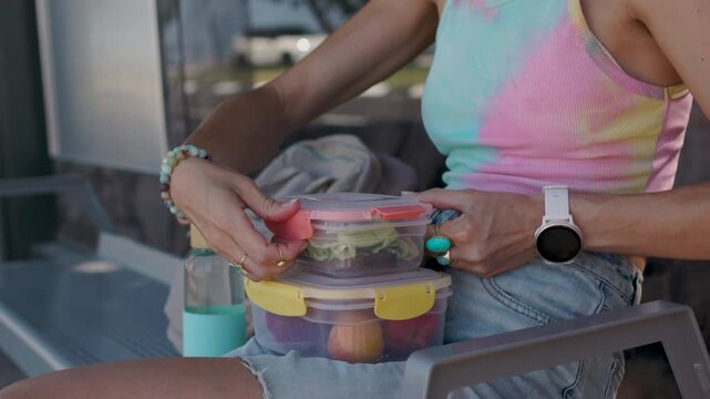 A young woman sits at a bus stop, holding a stack of clear lunch containers. She is waiting for the bus to work. Healthy eating at work. packed lunch.