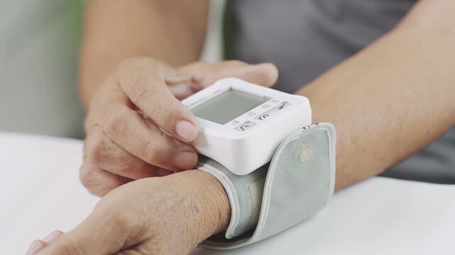 Close up of a senior using a digital wrist blood pressure monitor at home, pressing the start button for blood pressure measurement, representing hypertension management, selfcare.
