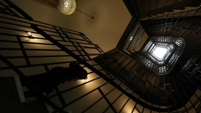 Silhouetted person descending multi-story stairwell with hand on banister, set inside rectangular shaft framed by concentric metal railings and stair treads that lead upward to bright white skylight