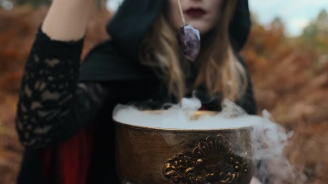 Close-up view shows ornate cauldron in foreground, thick smoke rising and spilling over rim while dark crystal pendant is held just above it by partially visible hooded figure in soft autumn light