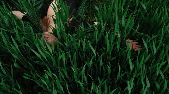High-angle view shows person lying face up within dense tall green grass, with light brown hair spread across blades, exposed shoulder, soft skin tones, and gentle movement through surrounding