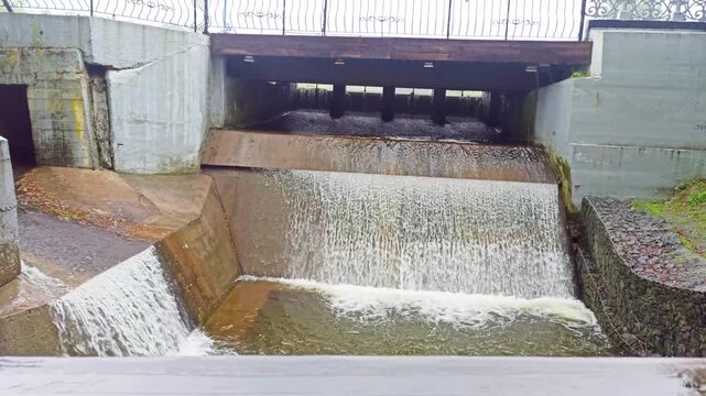 A small spillway under the bridge. Water flows through a concrete dam with a railing at the top, construction of a city canal, outdoor engineering, daytime.
