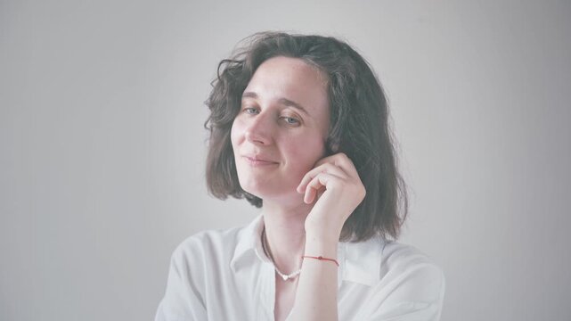 Young woman with curly hair in white shirt posing indoors, showing thoughtful and gentle smiles with hand near face, soft light and shallow depth of field.