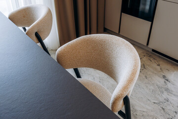Curved boucle dining chairs beside dark tabletop in modern kitchen with marble floor, neutral curtains, and minimalist cabinetry, creating soft refined interior styling © Kantemir Production
