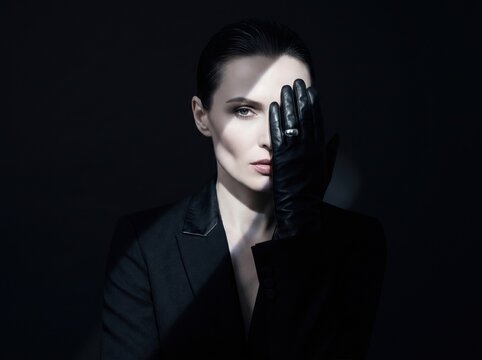 Woman covers half of her face with a black leather glove and stares into light and shadow. The composition features high-contrast moody lighting and minimal dark background.