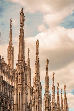 Multiple carved marble spires and statues on Milan Cathedral roof against clouds.