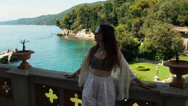 A young woman in sunglasses relaxes on a stone terrace by the sea in Trieste, with blue water, green gardens, coastal hills, and summer light around her.