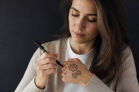 A young woman with long brown hair is writing or drawing on her own wrist in a dimly lit, dark setting.