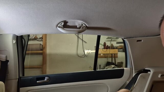 Car interior repair, man installs a grab handle onto the ceiling inside a vehicle. Static, eye-level medium shot.