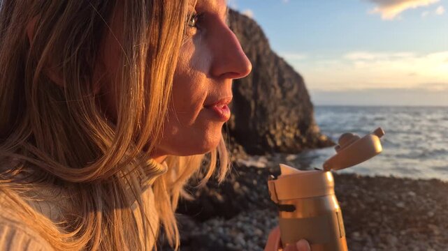 Woman eating snack, enjoying sunset at rocky seaside while holding portable thermos cup. Extreme close-up shot.
