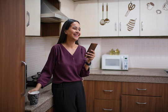 Smiling woman holding a cup of coffee and Smartphone in kitchen