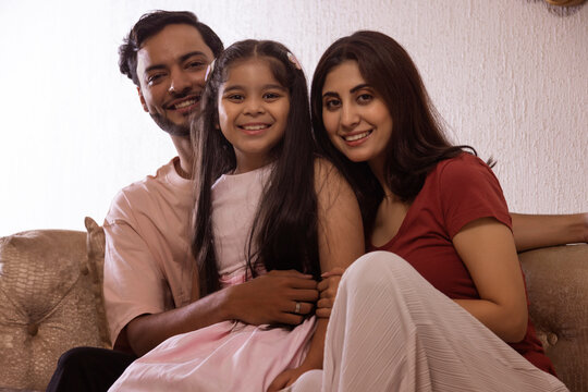 Young couple with their daughter looking at camera while sitting together on sofa at home