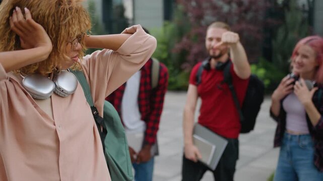 Group of students mocking humiliate bullying African American girl at college university outdoors multiracial classmates loud laugh violence discriminate sad female woman student stressed closing ears