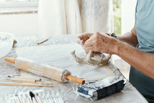 Male hands sculpting clay figure at table with tools in art studio. Creative handmade pottery, workshop and hobby concept.