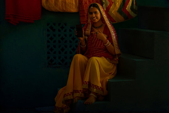 Portrait of Rajasthani woman pointing at mobile phone while sitting on stair at home