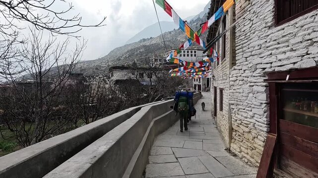 Trekker with backpack on narrow paved Marpha street, stone buildings prayer flags, Annapurna Circuit
