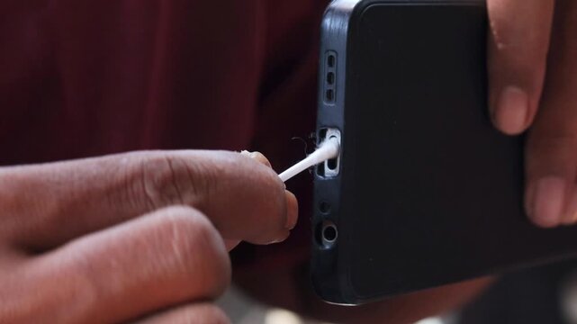 Close-up of an Asian man's hand cleaning a smartphone charging port with a cotton swab. Removing dust, lint, and debris from the charging connector for better device maintenance and smartphone care.