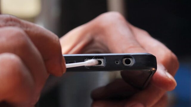 Close-up of an Asian man's hand cleaning a smartphone charging port with a cotton swab. Removing dust, lint, and debris from the charging connector for better device maintenance and smartphone care.