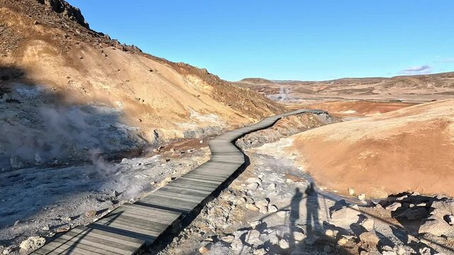 Seltun boardwalk winding through orange and ochre mineral hills with steam vents and human shadows, Reykjanes, Iceland