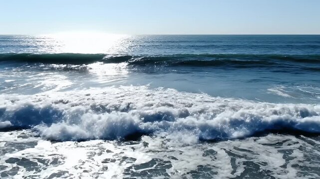 Sunlight glitters on the surface of the ocean as waves crash against the shore, creating a dynamic and refreshing coastal scene.